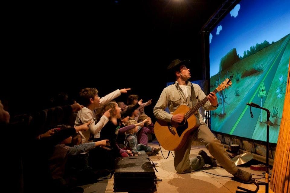 Un musicien jouant de la guitare devant un large écran de jeu, avec un public enthousiaste.